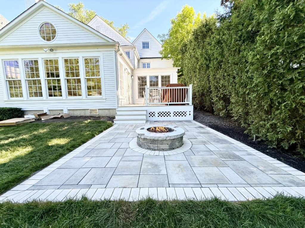 Gray paver patio with a round stone fire pit beside a white home